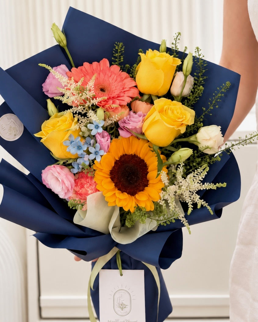 Close-up of Bright Futures graduation bouquet with golden sunflower, yellow roses, coral gerbera, soft pink blooms and delicate blue flowers.