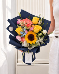 Colourful graduation bouquet featuring sunflower, yellow roses, pink lisianthus, white astilbe and blue oxypetalum in elegant navy wrapping.