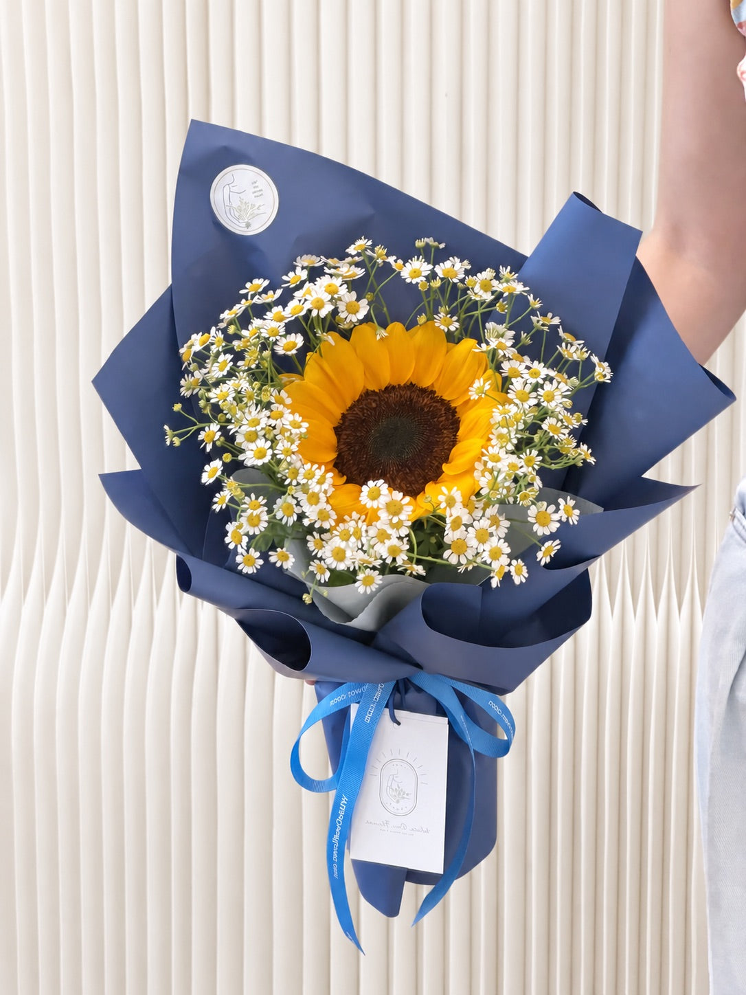 Little Sunshine graduation bouquet featuring one golden sunflower and white daisies, wrapped in navy blue paper with a bright blue ribbon.
